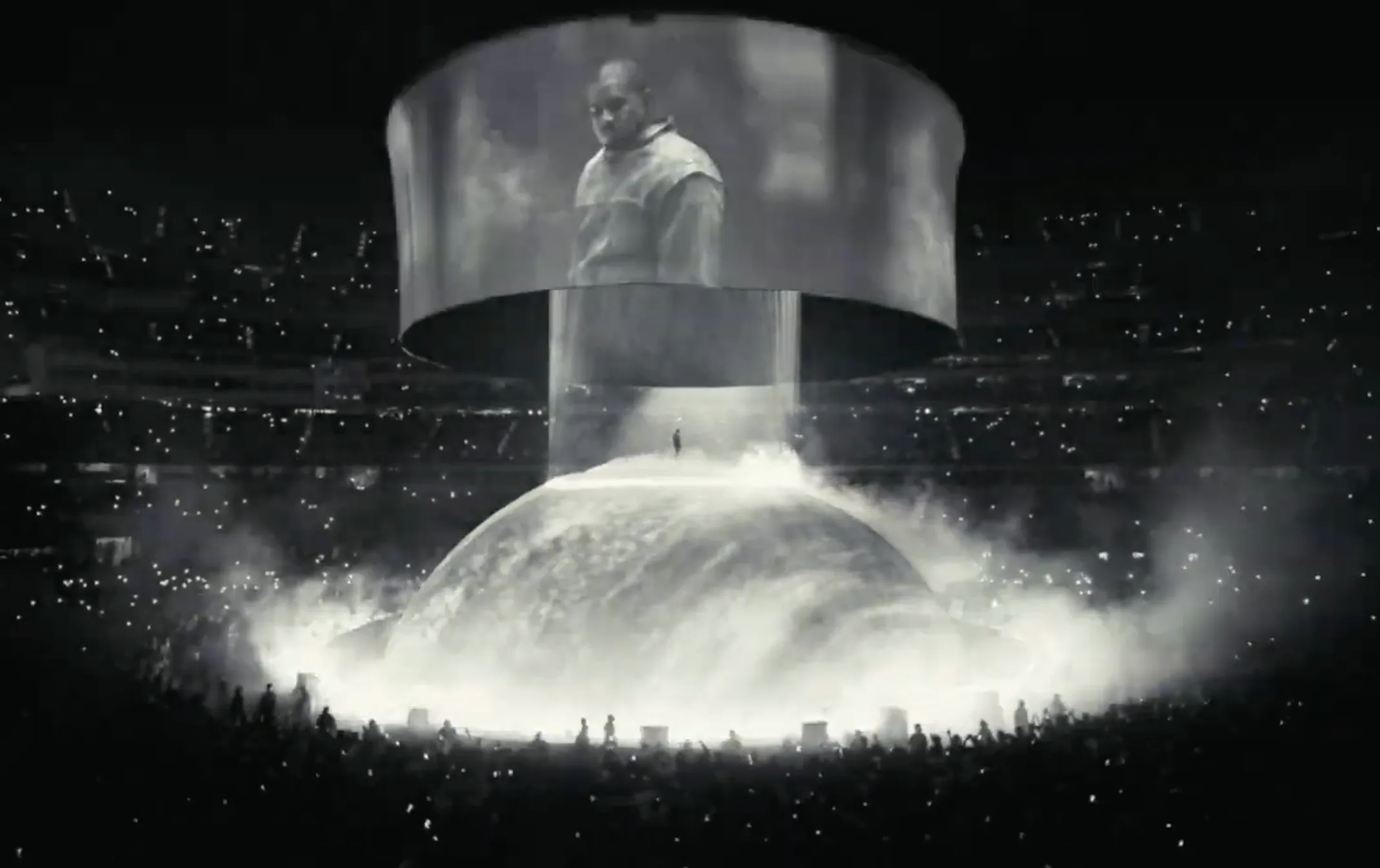 A stadium concert: a figure on a smoke-covered dome under a ring of screens
